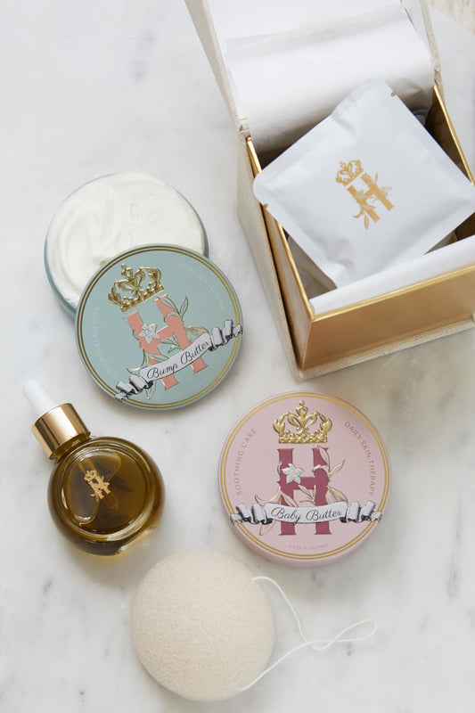 A luxury maternity skincare set arranged on a white marble surface. The set includes a small amber glass dropper bottle with Tender skin facial serum featuring a gold logo, two round decorative tin containers with whipped organic body butters, a beige box for white botanical soak sachets with gold logo laying on top of it, and a white konjac sponge.