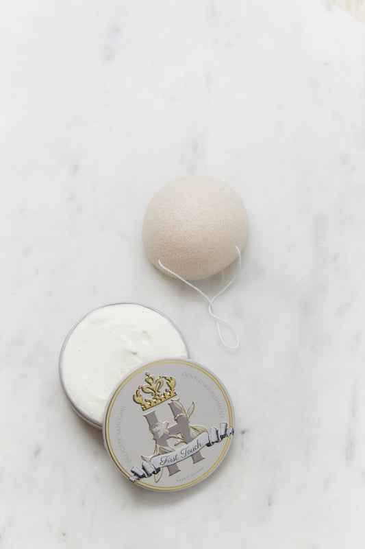 A round, off-white Konjac Sponge resting on a white marble surface next to its open warm grey tin, Royal Heir Botanicals First Touch butter, EWG verified, which features a decorative gold and grey crest label.