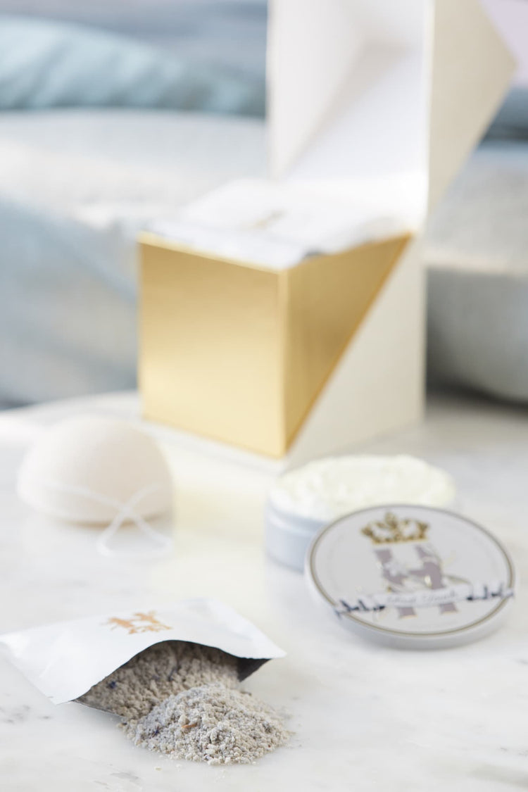 A luxury bath and body set featuring a white and gold geometric box, a white konjac sponge, a jar of white cream, a small round tin with Royal Heir Botanicals EWG verified First Touch butter with a crest, and a sachet with lavender botanical soak spilled on a white marble surface.