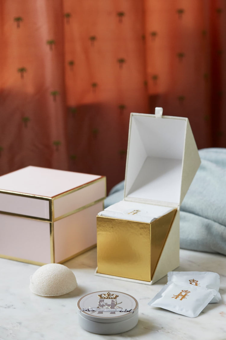 Pink gift box, an open gold box, a konjac sponge and a First Touch tin on a table with a blue blanket
