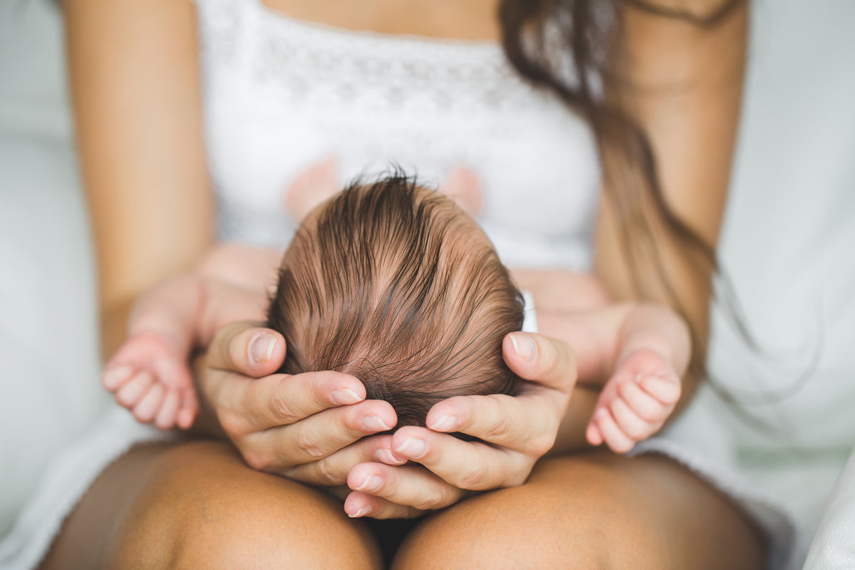 A close-up lifestyle shot of a mother holding the head of her newborn baby, with her hands cradling the infant's head and small hands visible on both sides.