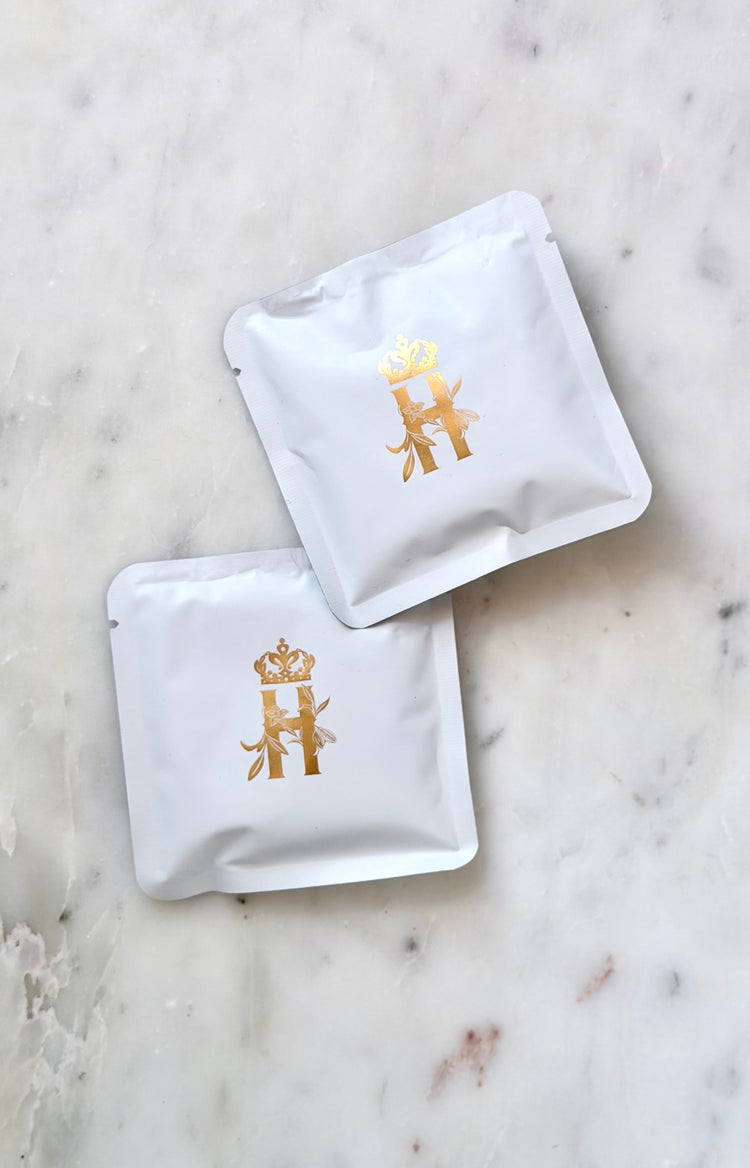 Two small white Royal Heir Botanicals soak sachets with a gold crowned H, on white marble