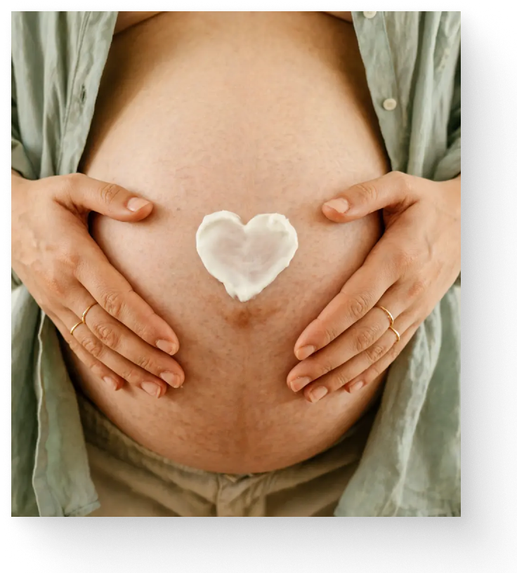 A close-up of a pregnant woman's bare belly with both hands resting on it, wearing an open sage green shirt. A small white heart-shaped cosmetic butter is placed on the center of the belly.