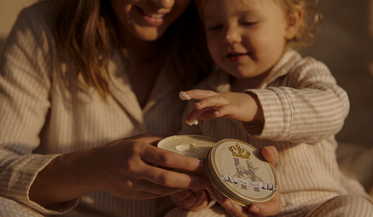 warm scene of a mother with her toddler sitting on her lap dipping their finger into a freshly opened Royal Heir Botanicals First Touch whipped organic Butter. The mom and baby are wearing matching white and cream striped pajamas. 