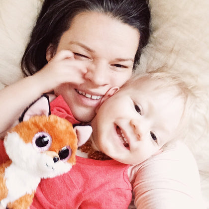 A close-up lifestyle shot of a smiling woman and a baby lying on a soft cream-colored blanket, with the woman holding a plush orange and white stuffed fox.