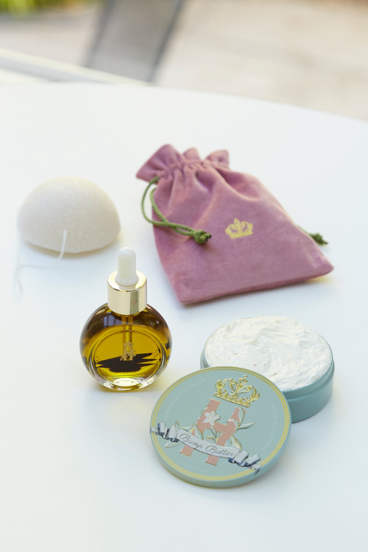 Pink velvet pouch, a serum bottle, a konjac sponge and a Bump Butter tin on a white table