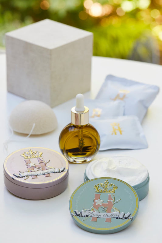 A luxury maternity skincare set arranged on a white surface with a blurred green outdoor background. The set includes a small amber glass dropper bottle with Tender skin facial serum, two round decorative tin containers with whipped organic body butters, a beige box for white botanical soak sachets with gold logo laying next to it, and a white konjac sponge.