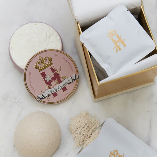 Open gold-lined box with a Butter in Pink tin, an open tin of butter, soak sachets, a konjac sponge and spilled soak powder