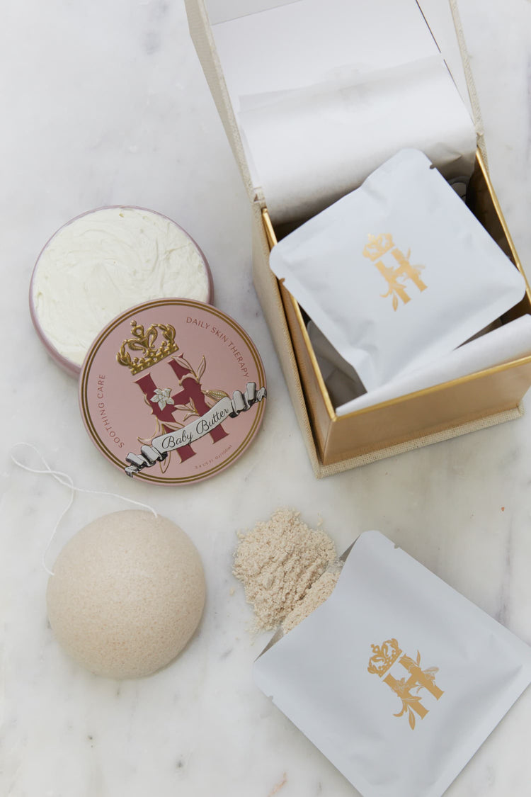 Open gold-lined box with a Butter in Pink tin, an open tin of butter, soak sachets, a konjac sponge and spilled soak powder