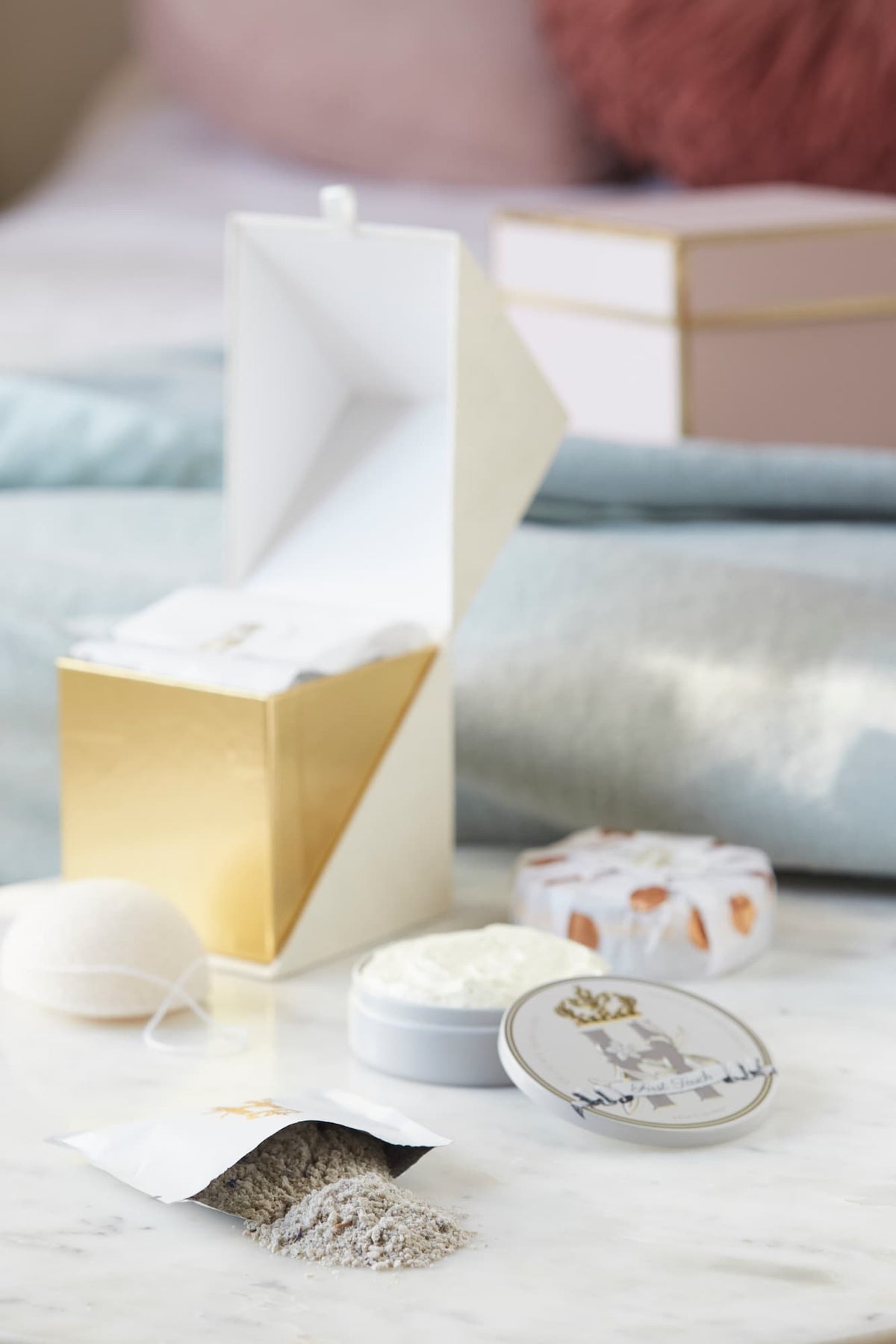 Luxury skincare set on white marble surface with blurred pastel pink and blue  background featuring a cream and gold cube shaped box open with a konjac sponge with a string, round tin with a crest logo and a gold crown holding Royal Heir Botanicals whipped organic, EWG verified First Touch Butter for very delicate skin and a couple of white sachets with lavender botanical soak and a gold Royal Heir logo.