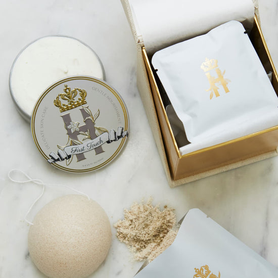 Luxury skincare set on white marble surface  featuring a cream and gold cube shaped box open with a konjac sponge with a string, round tin with a crest logo and a gold crown holding Royal Heir Botanicals whipped organic, EWG verified First Touch Butter for very delicate skin and a couple of white sachets with lavender botanical soak and a gold Royal Heir logo.