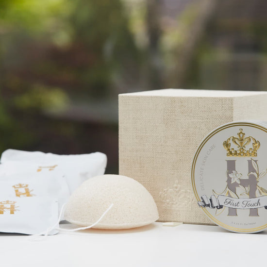 Luxury skincare set on white surface with blurred green foliage background featuring a cream and gold cube shaped box open with a konjac sponge with a string, round tin with a crest logo and a gold crown holding Royal Heir Botanicals whipped organic, EWG verified First Touch Butter for very delicate skin and a couple of white sachets with lavender botanical soak and a gold Royal Heir logo.