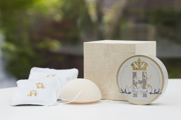 Luxury skincare set on white surface with blurred green foliage background featuring a cream and gold cube shaped box open with a konjac sponge with a string, round tin with a crest logo and a gold crown holding Royal Heir Botanicals whipped organic, EWG verified First Touch Butter for very delicate skin and a couple of white sachets with lavender botanical soak and a gold Royal Heir logo.