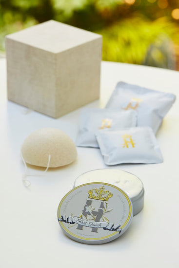 Luxury skincare set on white surface with blurred green foliage background featuring a cream and gold cube shaped box with a konjac sponge with a string, round tin with a crest logo and a gold crown holding Royal Heir Botanicals whipped organic, EWG verified First Touch Butter for very delicate skin and a couple of white sachets with lavender botanical soak and a gold Royal Heir logo.