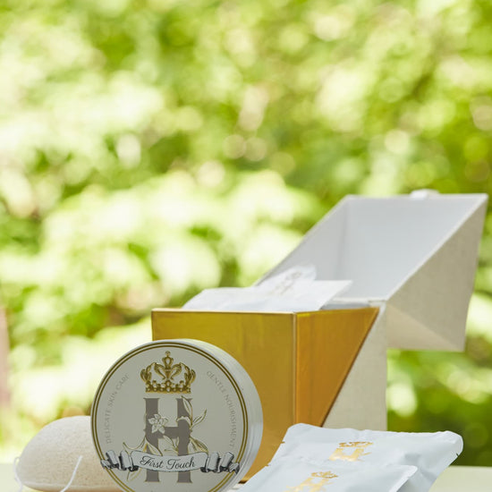 Luxury skincare set on white surface with blurred green foliage background featuring a cream and gold cube shaped box open with a konjac sponge with a string, round tin with a crest logo and a gold crown holding Royal Heir Botanicals whipped organic, EWG verified First Touch Butter for very delicate skin and a couple of white sachets with lavender botanical soak and a gold Royal Heir logo.