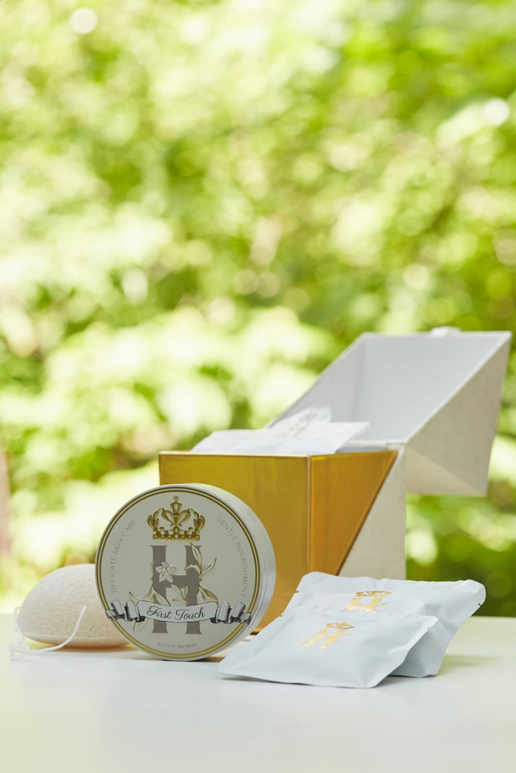 Luxury skincare set on white surface with blurred green foliage background featuring a cream and gold cube shaped box open with a konjac sponge with a string, round tin with a crest logo and a gold crown holding Royal Heir Botanicals whipped organic, EWG verified First Touch Butter for very delicate skin and a couple of white sachets with lavender botanical soak and a gold Royal Heir logo.