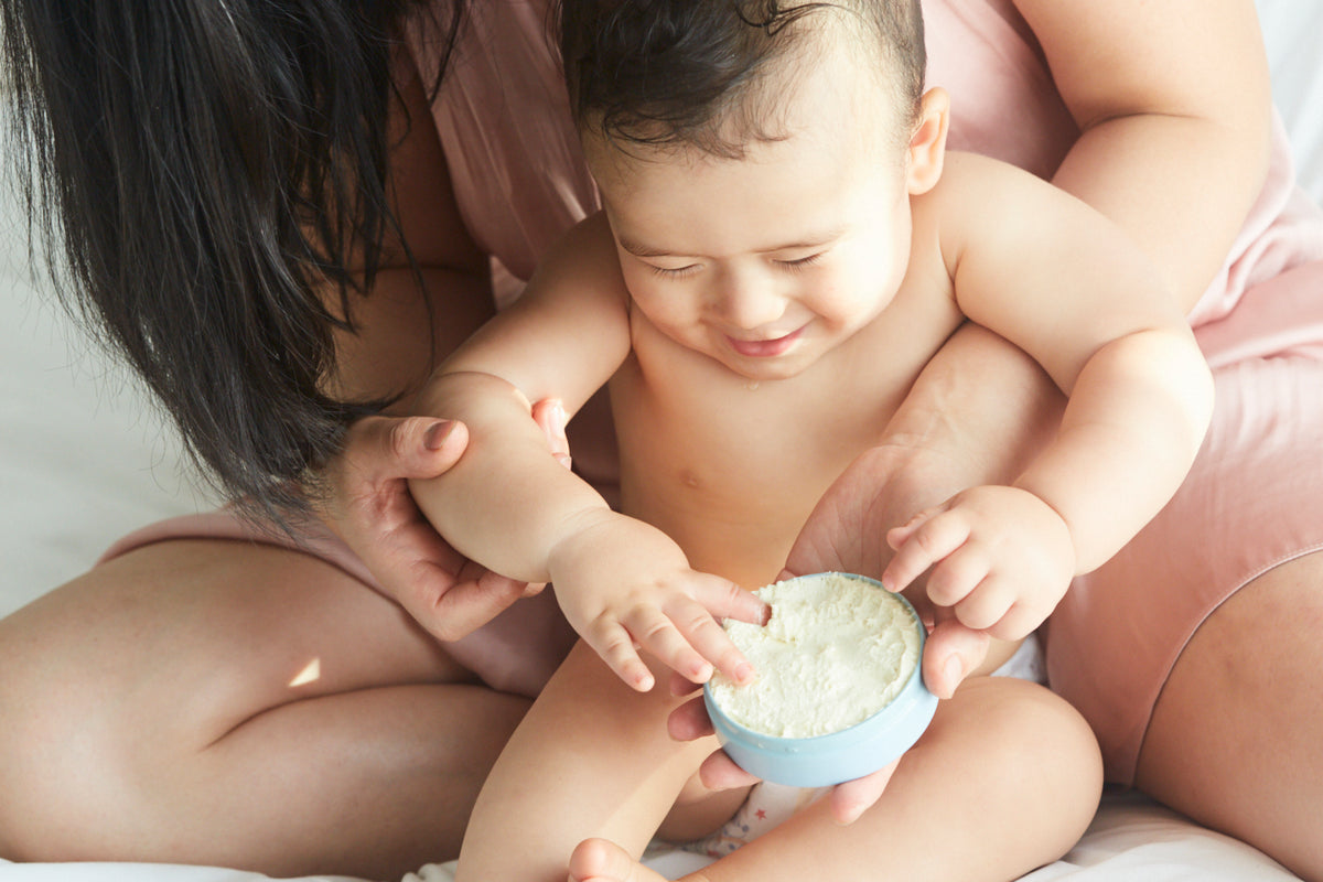 Mother and her toddlersitting n a bed facing the camera with a blue tin with butter in mother's hand and baby joyfully reaching in 