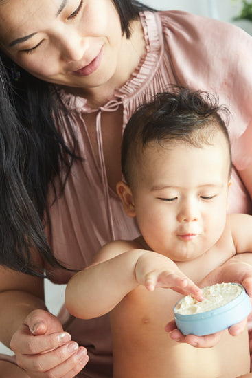 Mother holding her baby, who reaches into an open tin of Butter in Blue