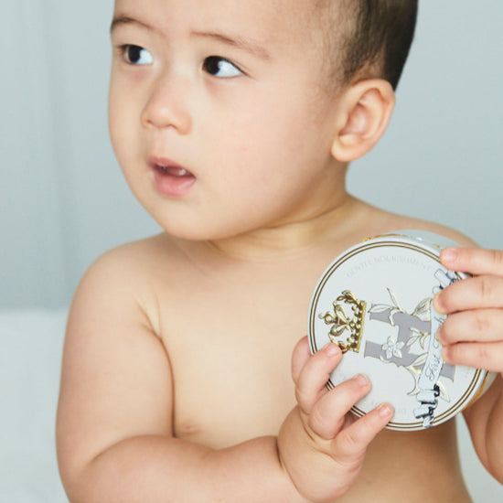 Baby holding a tin of Royal Heir Botanicals First Touch EWG verified Cocoa butter based butter for babies from day 1, the next best thing to Vernix