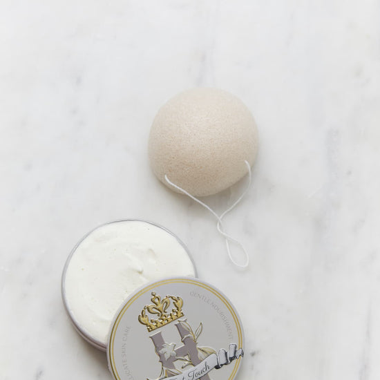 A round, off-white Konjac Sponge resting on a white marble surface next to its open warm grey tin, Royal Heir Botanicals First Touch butter, EWG verified, which features a decorative gold and grey crest label.
