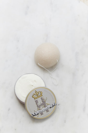 A round, off-white Konjac Sponge resting on a white marble surface next to its open warm grey tin, Royal Heir Botanicals First Touch butter, EWG verified, which features a decorative gold and grey crest label.