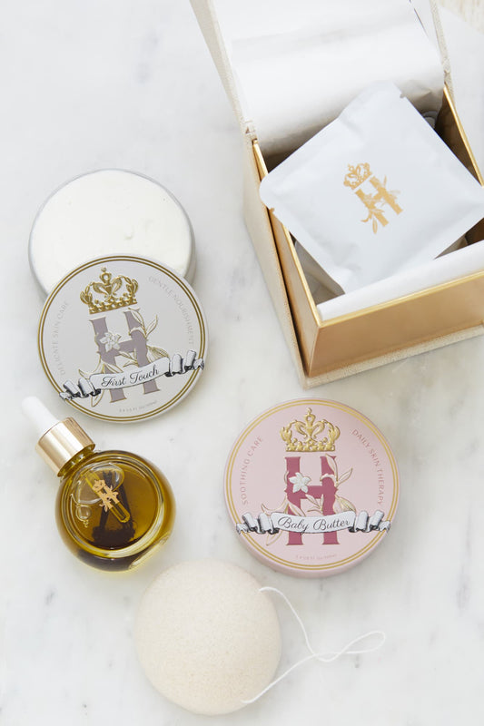 A luxury skincare gift set for a new mother and a baby by Royal Heir Botanicals arranged on a white marble surface, featuring a gold-toned box with a white botanical soak sachet, a round tin labeled 'Baby Butter in Pink', and a tin with First Touch butter, a small dropper bottle with facial serum, konjac exfoliating sponge.
