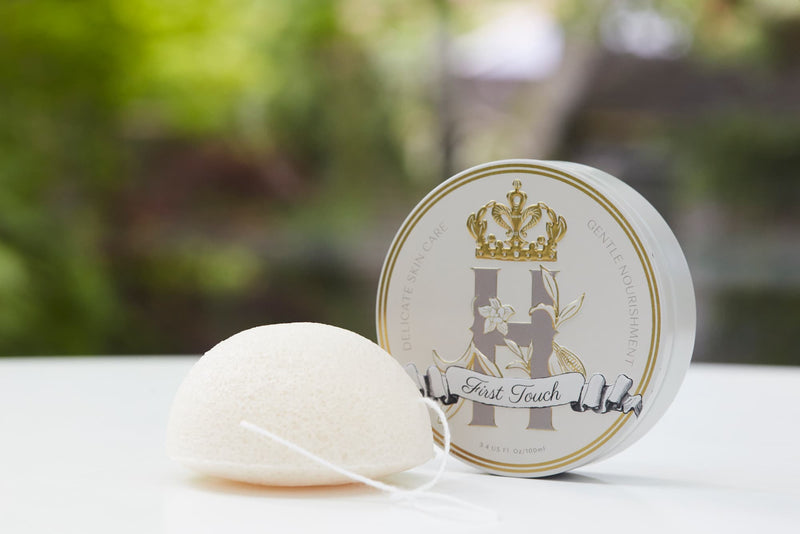 A round white Konjac  sponge with a white string loop sits in front of a round white and gold branded tin with Royal Heir Botanicals First Touch butter, EWG verified on a white surface with a blurred green outdoor background. 