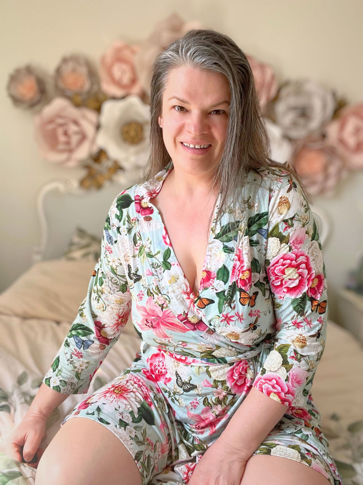 Henrieta, founder of Royal Heir Botanicals, smiling in a floral wrap dress in front of a wall of paper flowers.