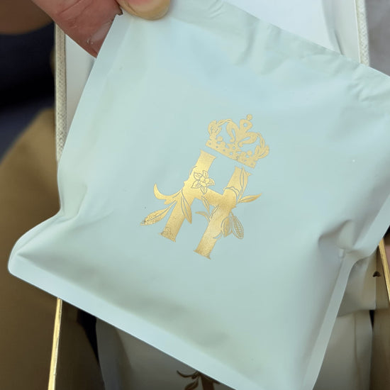 Hand lifting a sage-green soak sachet stamped with a gold H from an open box