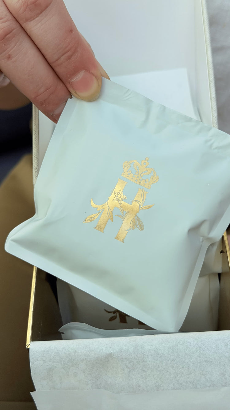 Hand lifting a sage-green soak sachet stamped with a gold H from an open box
