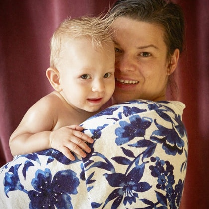 A baby wrapped in a cream-colored towel with a bold blue floral pattern, held by a smiling woman against a dark red curtain background.