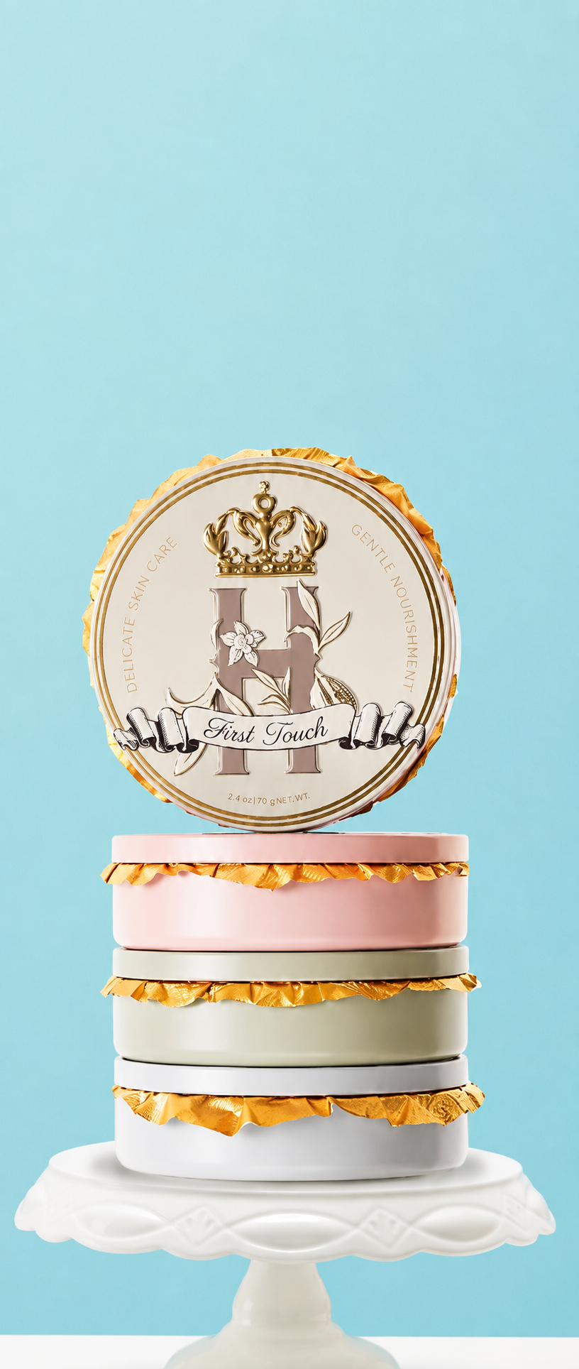 A decorative cake -like stack of Royal Heir Botanicals whipped butters on a white pedestal stand against a light blue background. The cake has three stacked tiers in white, pink, and gray with gold ruffled trim, topped with a round gold and white cake topper featuring a crown and the words 'First Touch'. EWG verified organic butter for newborn babies. 