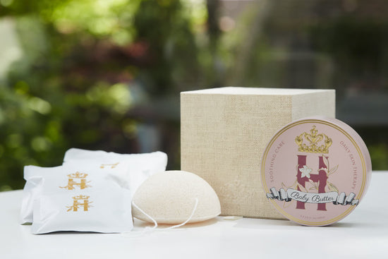 Luxury maternity skincare set featuring a pink round decorative tin with organic whipped body butter, cream colored cube box, white konjac sponge with a string and a few white sachets with rose botanical soak with golden Royal Heir Botanicals Logo