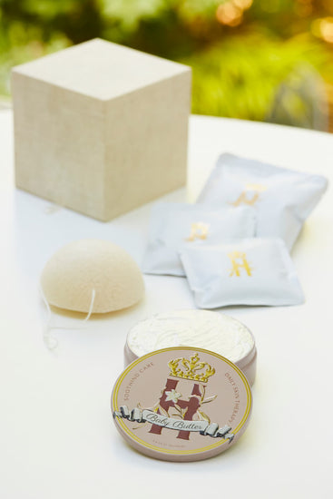 Luxury maternity skincare set featuring a pink round decorative tin with organic whipped body butter, cream colored cube box, white konjac sponge with a string and a few white sachets with rose botanical soak with golden Royal Heir Botanicals Logo