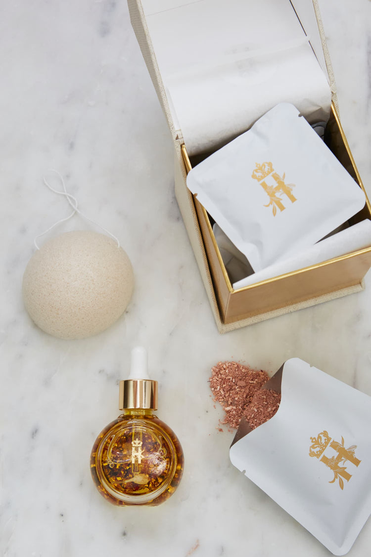 Open box with soak sachets, a konjac sponge, a serum bottle with rose petals and spilled soak powder on marble