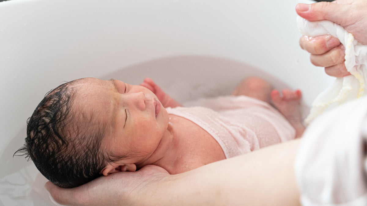 Newborn baby being bathed with a white towel draped over the side of the tub.