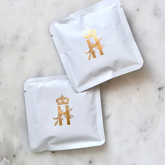 Two small white Royal Heir Botanicals soak sachets with a gold crowned H, on white marble