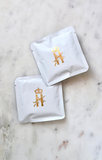 Two small white Royal Heir Botanicals soak sachets with a gold crowned H, on white marble