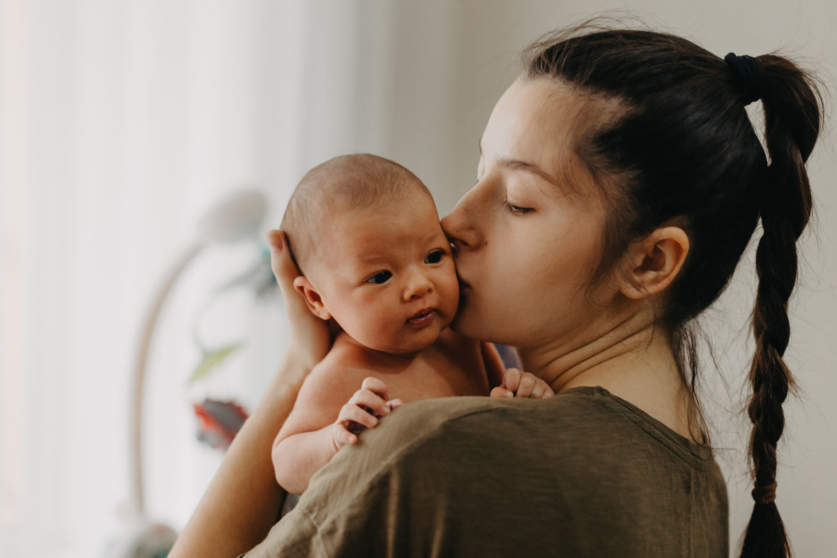 A close-up lifestyle shot of a woman with a dark braided ponytail kissing a baby on the cheek while holding the baby against her chest. The woman is wearing a muted olive-brown top, and the background is a soft, brightly lit, neutral indoor setting.