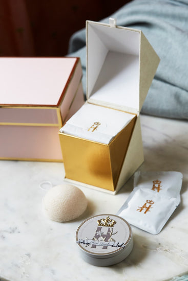 Open pink and gold gift box with a soak sachet, a konjac sponge and a First Touch tin