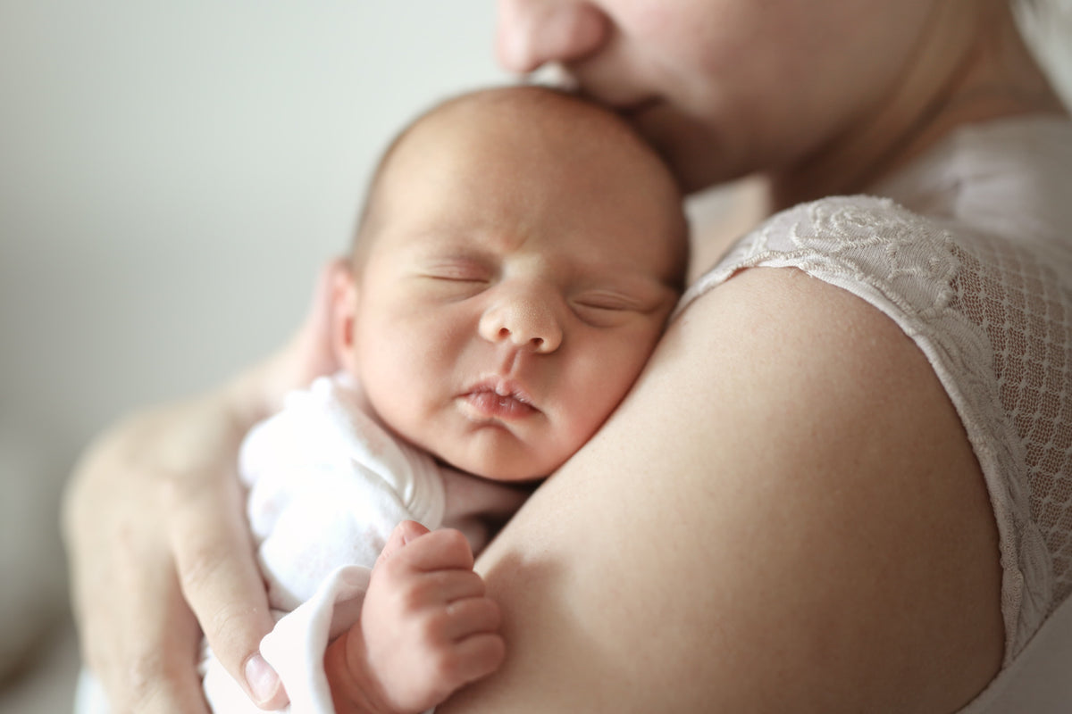 A close-up lifestyle shot of a sleeping newborn baby nestled in the shoulder of a mother, wearing a white onesie and a white lace-trimmed top.