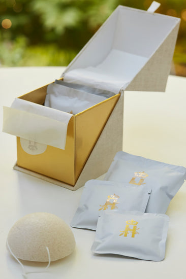 Open gold ritual box with soak sachets and a konjac sponge on a table outdoors