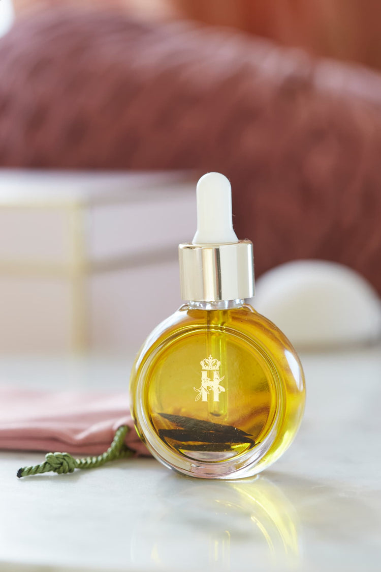 A round glass dropper bottle filled with golden yellow Tender skin facial serum by Royal Heir Botanicals, featuring a gold cap and a white rubber pipette. The bottle has a gold monogram logo on the front and is set on a white marble surface with a pink fabric and a blurred brown background.