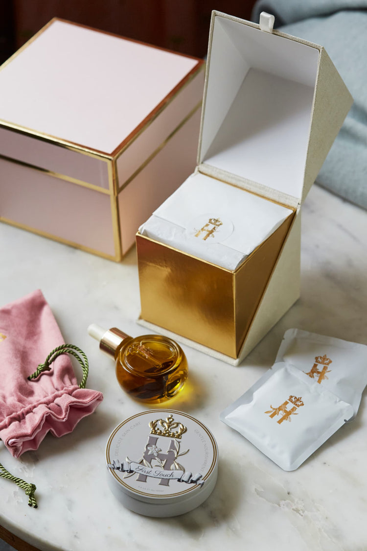 Open pink gift box beside a serum bottle, a pink pouch, a First Touch tin and soak sachets