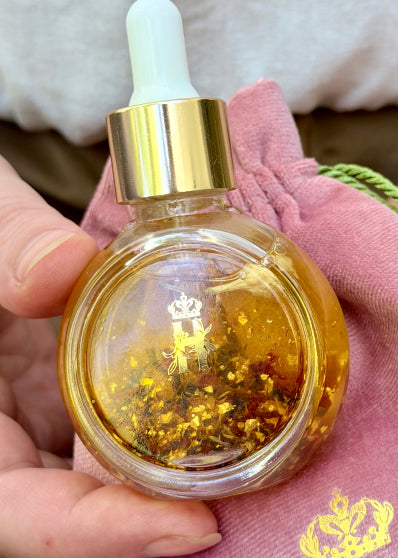 A hand holds a small round glass dropper bottle filled with golden amber Balancing Rose Facial serum and embedded gold flakes, with a pink drawstring pouch and a little gold crown logo behind it