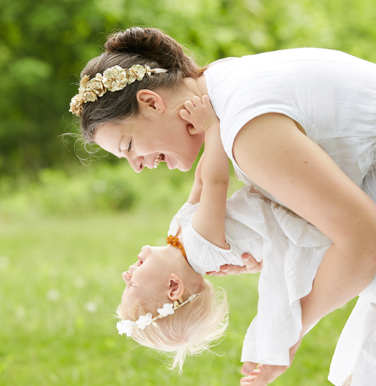 A woman in a white sleeveless dress and a baby in a matching white outfit play together outdoors on a grassy lawn. The woman wears a gold floral headband, and the baby wears a small white flower headband.