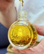 A hand holds a small round glass dropper bottle filled with golden amber Balancing  Rose Facial serum and embedded gold flakes, dripping some of the oil into the bottle from a dropper