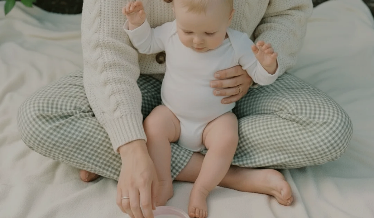 A baby in a white long-sleeve bodysuit sits on a mother's lap wearing a cream knit sweater and green-and-white gingham pants, seated on a light fabric surface, reaching for Royal Heir Botanicals baby butter in Pink
