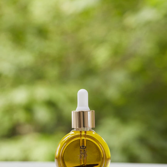 Tender Skin Serum in a round glass bottle with a gold dropper, in front of green foliage