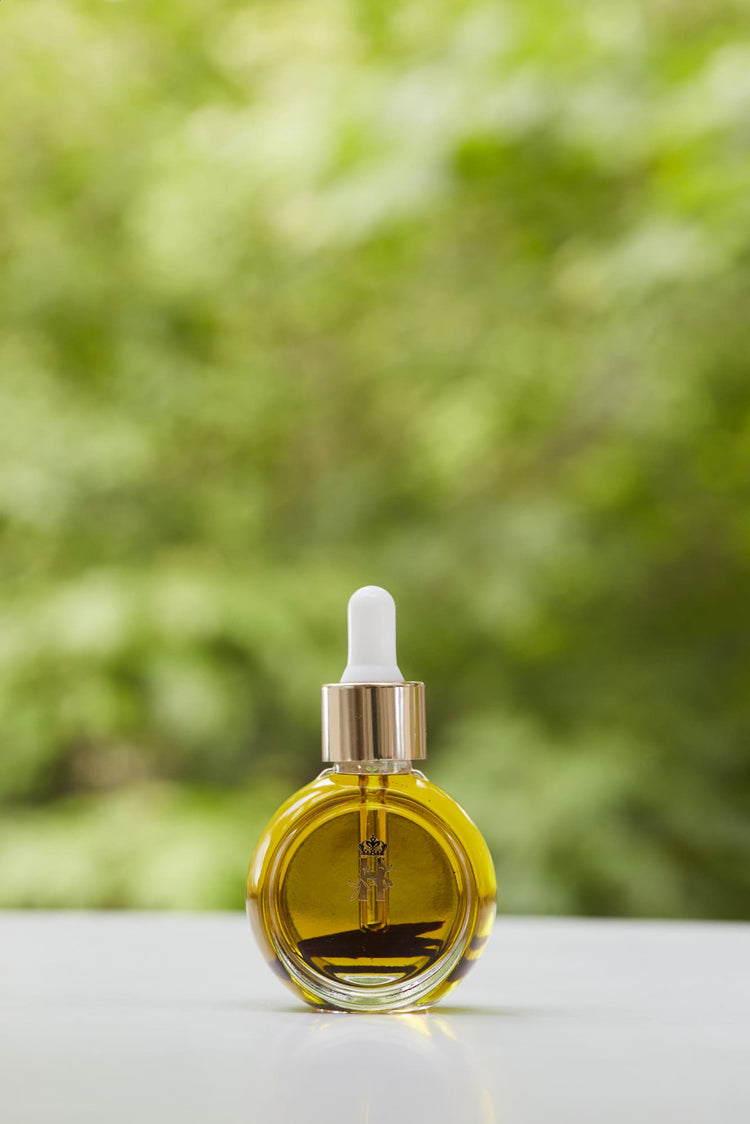 Tender Skin Serum in a round glass bottle with a gold dropper, in front of green foliage